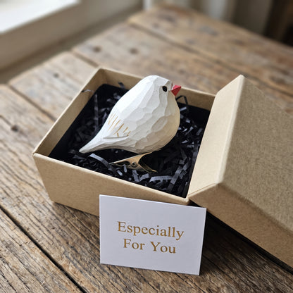 A white geometric Zebra Finch ornament with a red beak resting in black shredded paper inside a kraft gift box, with a small card reading 'Especially For You' on a rustic wooden table.