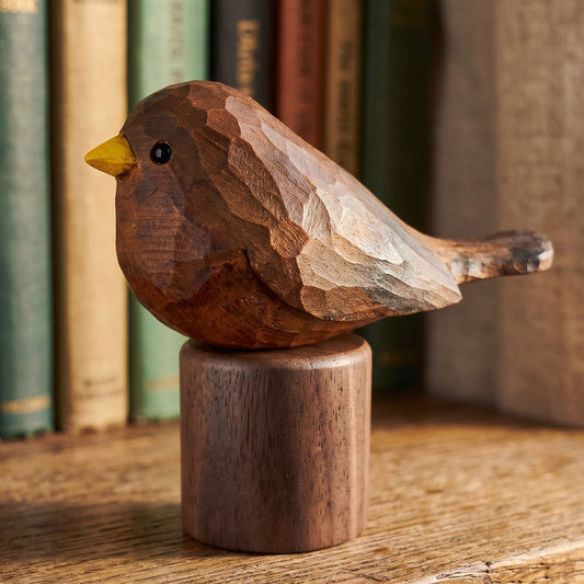 A carved wooden Eurasian Blackbird figurine with a yellow beak and black eye, mounted on a cylindrical wooden base and displayed on a wooden shelf in front of a blurred background of books.