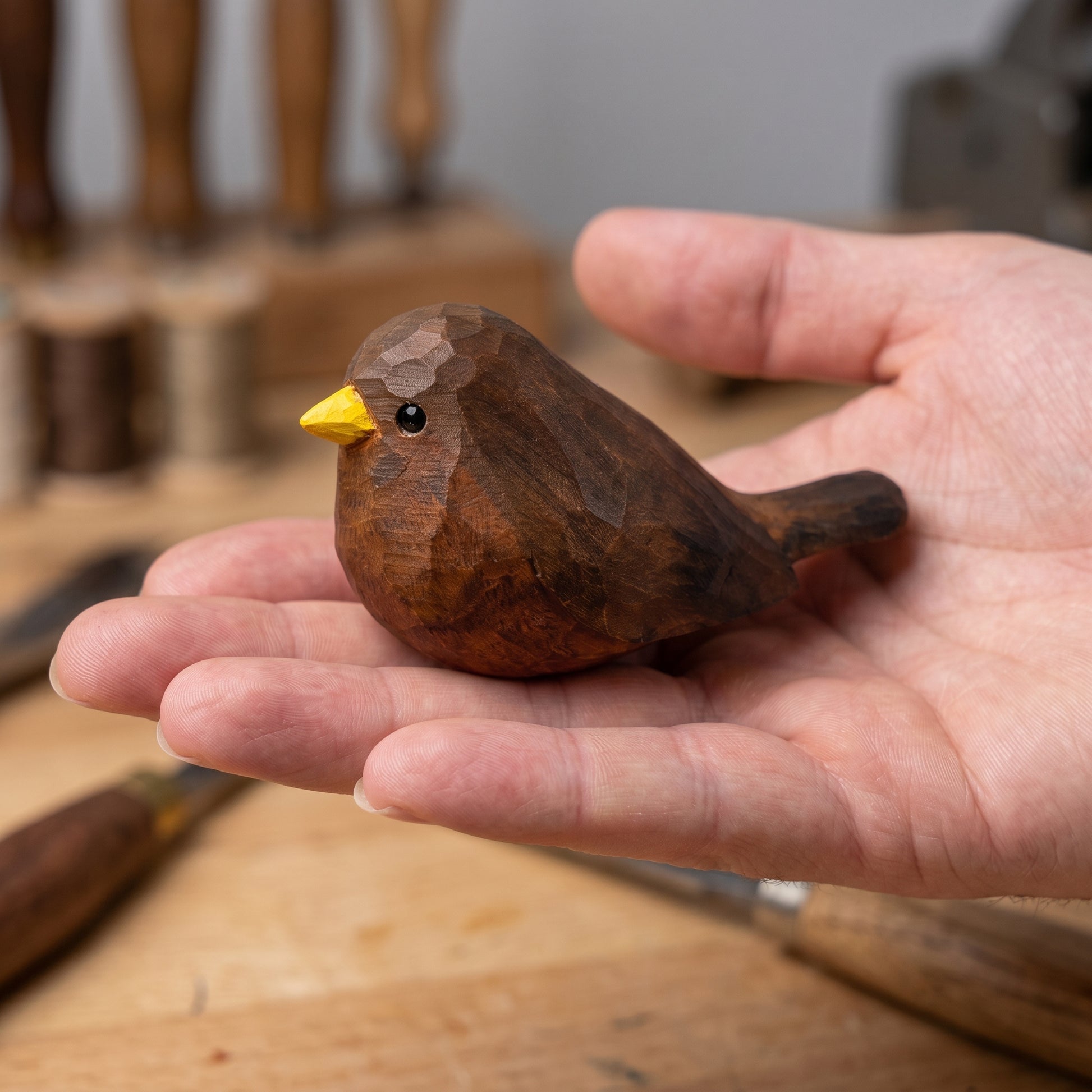A hand-carved wooden Eurasian Blackbird figurine with a bright yellow beak, held gently in a person's hand over a wooden workbench.