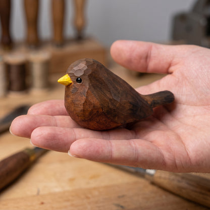 A hand-carved wooden Eurasian Blackbird figurine with a bright yellow beak, held gently in a person's hand over a wooden workbench.