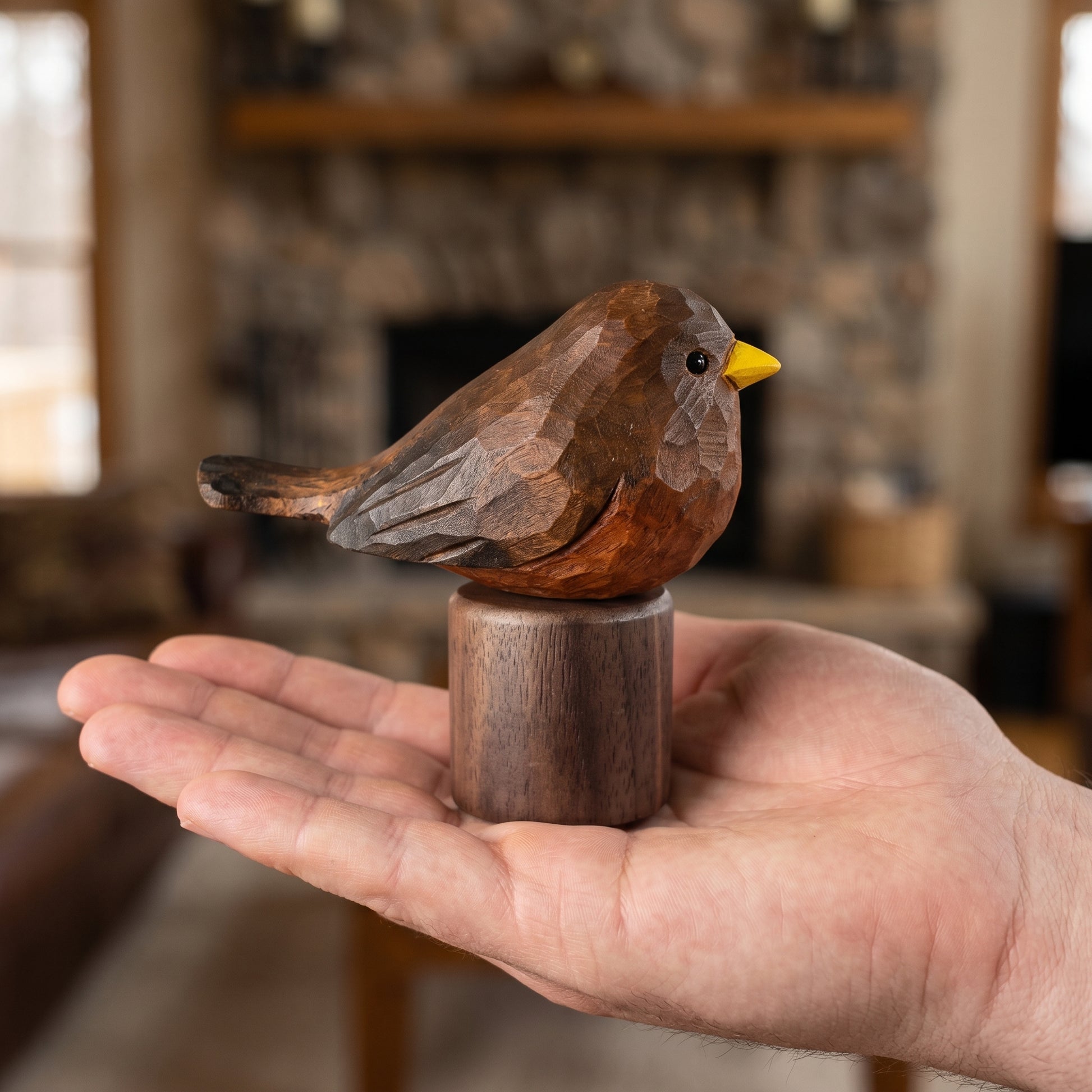 A hand holds a small carved wooden Eurasian Blackbird sculpture on a cylindrical wooden base, with a blurred indoor fireplace background.