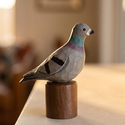 A handcrafted wooden Rock dove sculpture of a pigeon with painted gray, black, and iridescent green accents, mounted on a cylindrical wooden base and photographed on a tabletop in a softly blurred indoor setting.