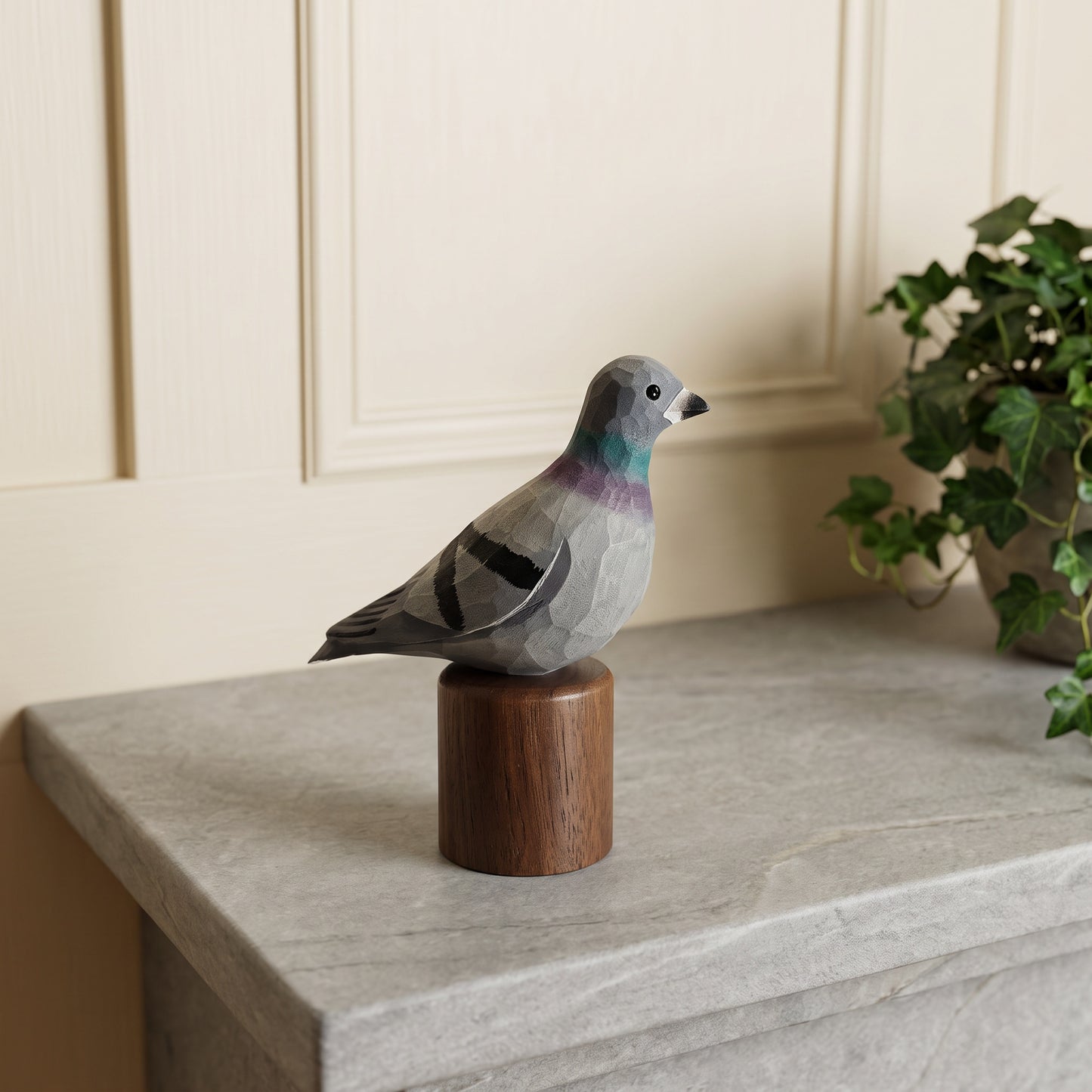 A handcrafted wooden Rock dove sculpture of a pigeon, mounted on a cylindrical wooden base, displayed on a gray stone ledge with a potted ivy plant in the background.