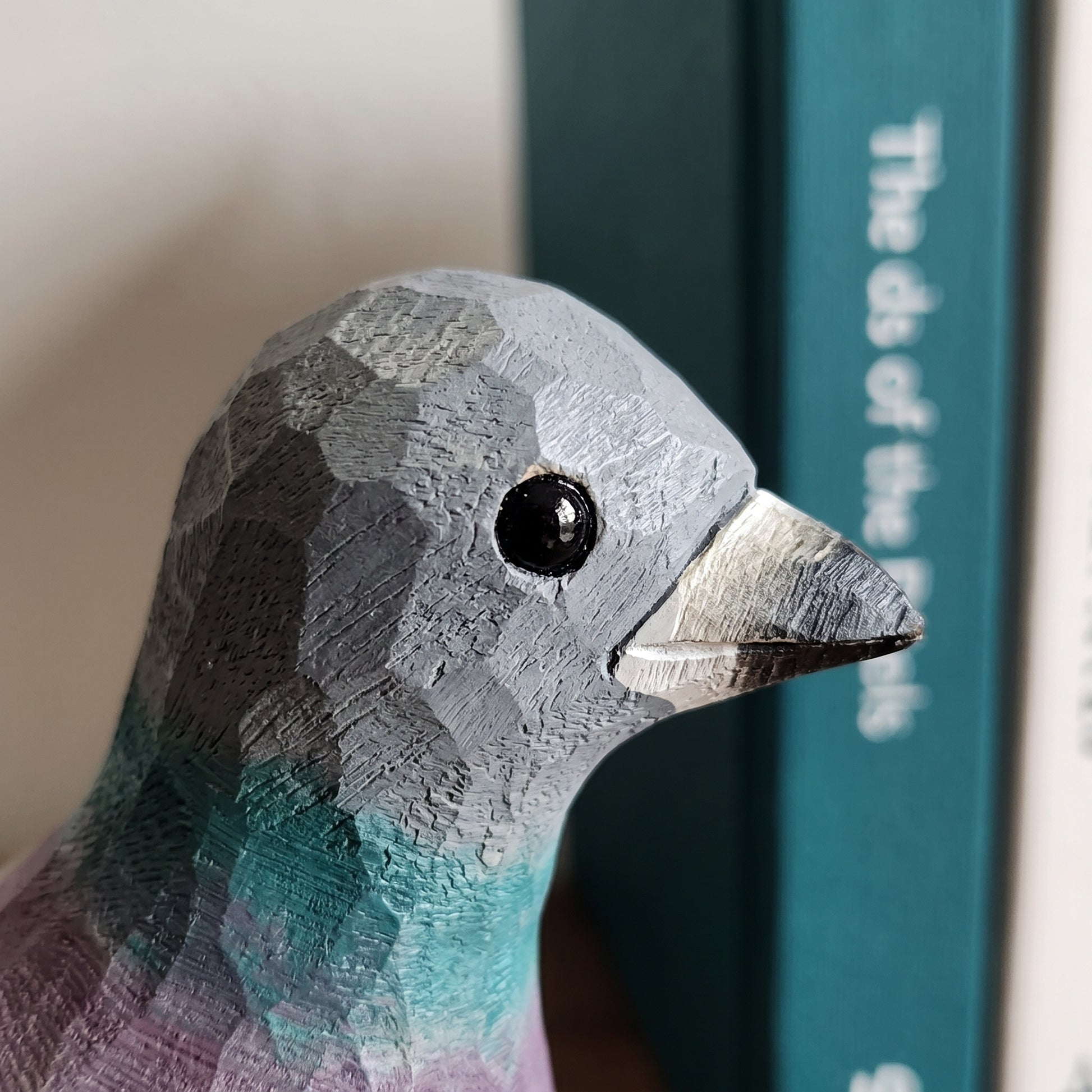 Close-up of a carved Rock dove figurine with a geometric, faceted texture. The bird has a gray head, a teal and purple neck, and a metallic silver beak.