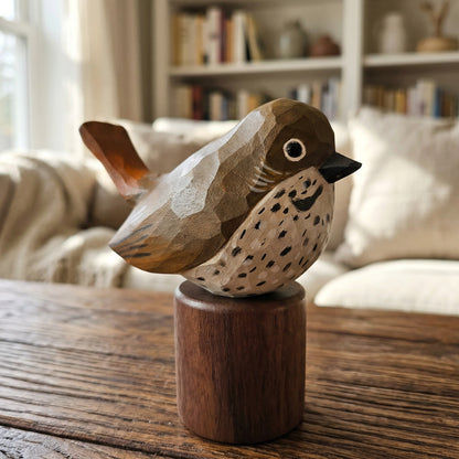 A hand-carved wooden Hermit Thrush figurine with a natural wood base, displayed on a wooden table in a cozy, softly blurred living room setting.