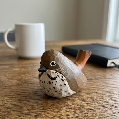 A small hand-carved wooden Hermit Thrush figurine with a brown and beige speckled body, placed on a wooden table with a blurred white mug and a black notebook in the background.