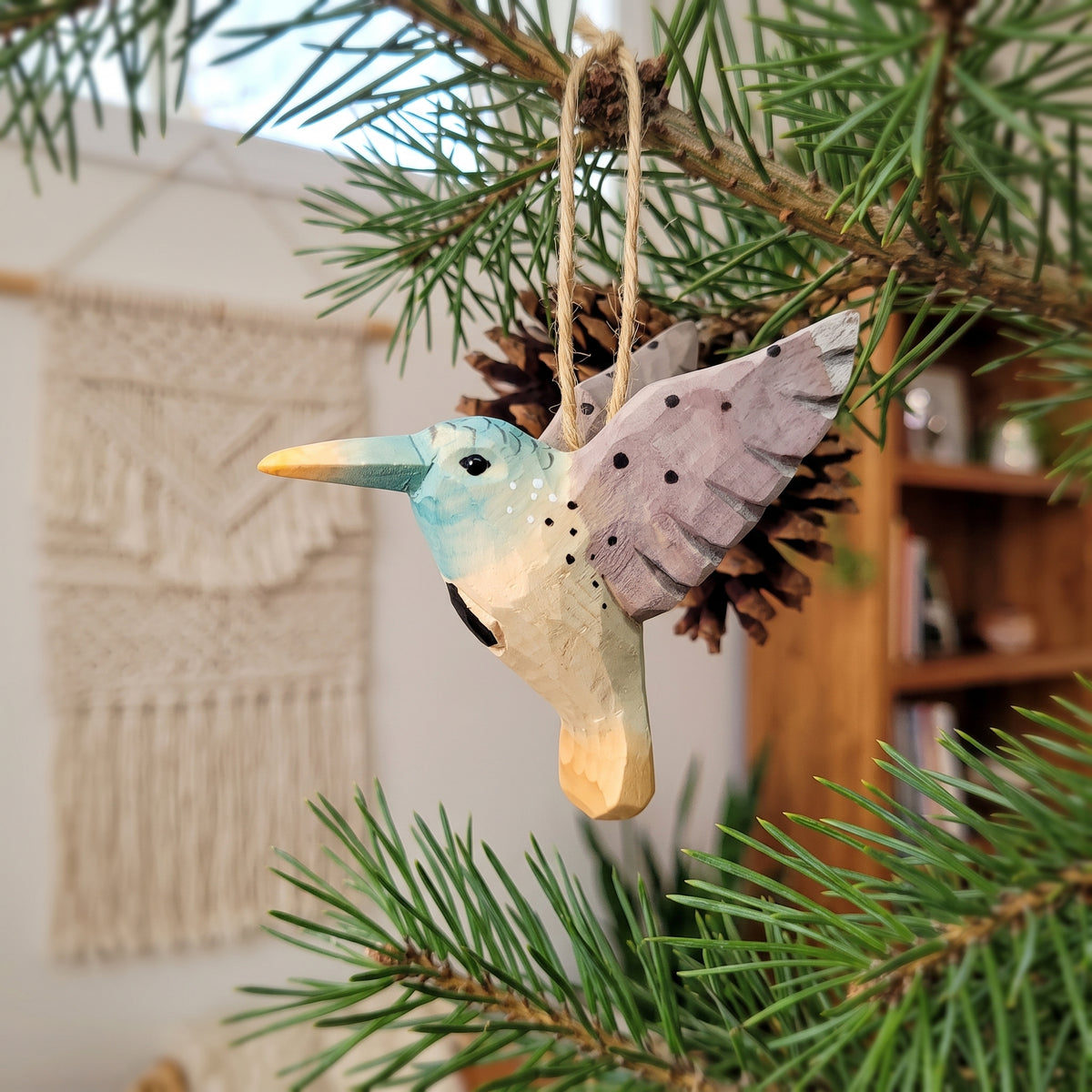 Hand-carved wooden hummingbird ornament with light blue, beige, and speckled gray wings hanging from a pine branch.