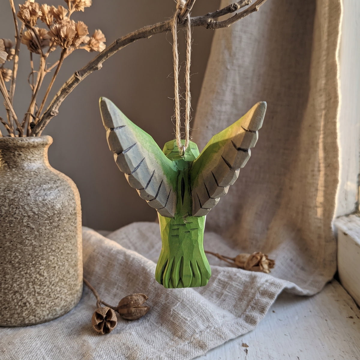 Green hand-carved basswood hummingbird hanging ornament displayed in a rustic window setting with dried flowers.