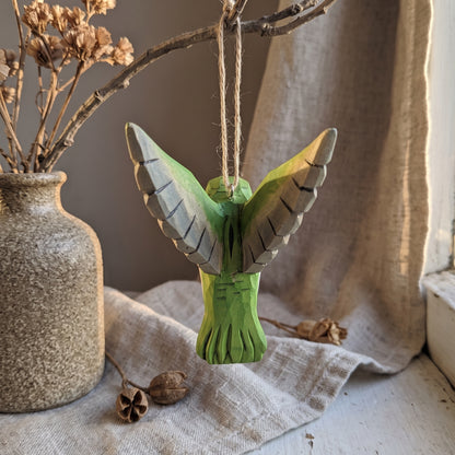 Green hand-carved basswood hummingbird hanging ornament displayed in a rustic window setting with dried flowers.