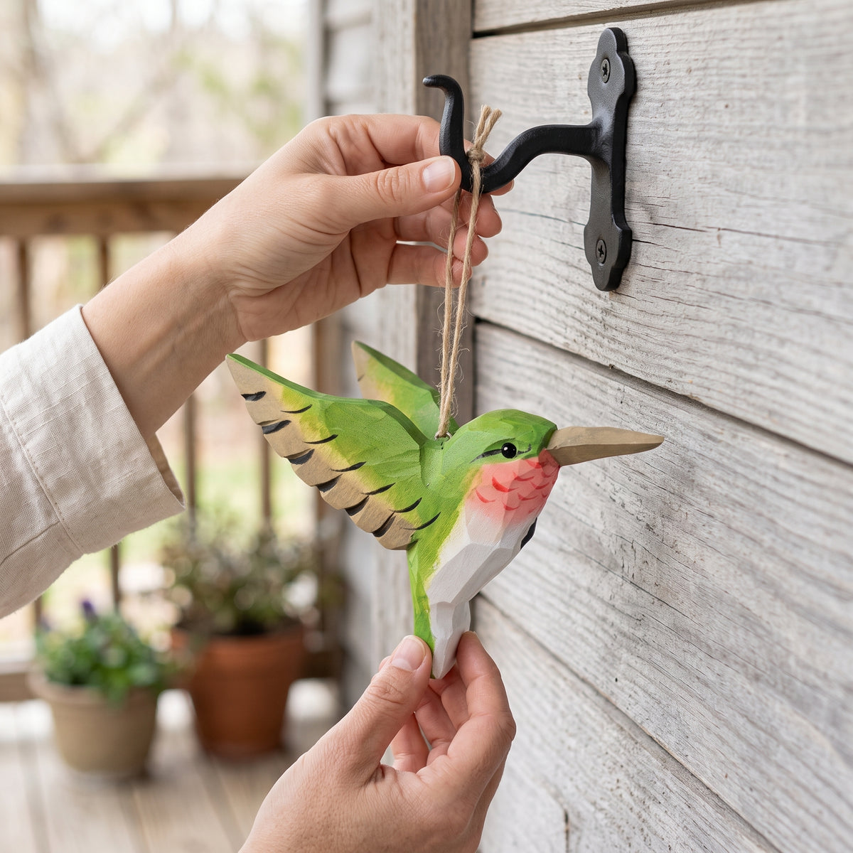 Green hand-carved basswood hummingbird hanging ornament displayed on a wrought-iron hook on a porch wall.