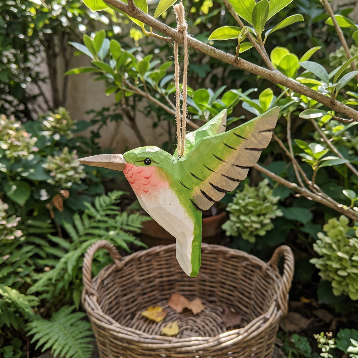 Green hand-carved basswood hummingbird hanging ornament displayed outdoors in a garden with ferns and a wicker basket.