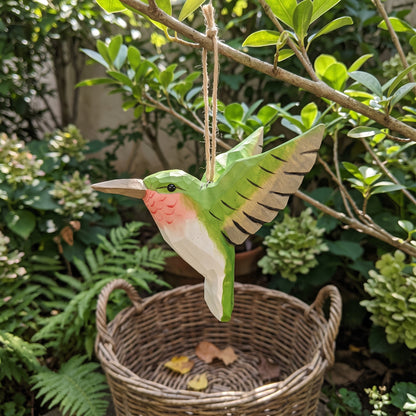 Green hand-carved basswood hummingbird hanging ornament displayed outdoors in a garden with ferns and a wicker basket.