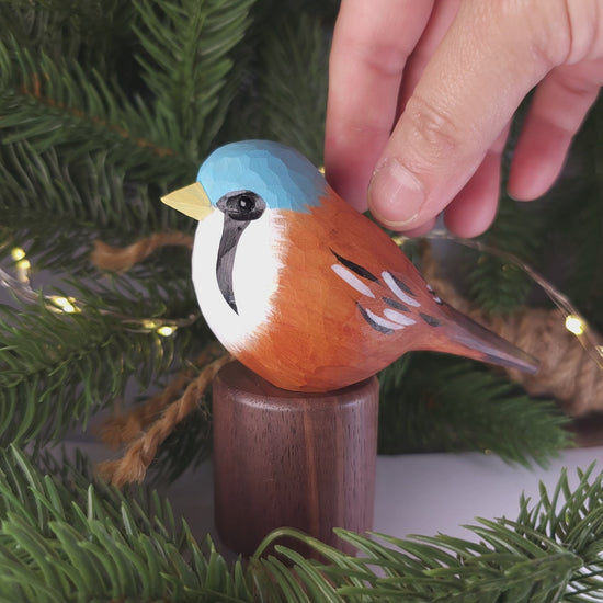 Hand-carved and painted wooden Bearded Tit figurine mounted on a dark wood base set against pine branches and warm fairy lights