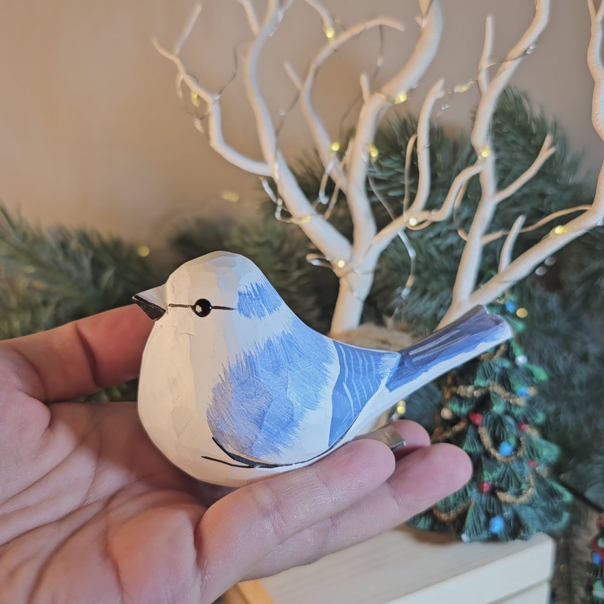 A close-up side-profile shot of a hand-carved wooden Azure Tit clip-on ornament held in an open hand, featuring a white head with a dark eye-stripe, light blue upperparts with textured white wing bars, a blue tail, and a silver metal clip attached underneath, set against a softly blurred background with white decorative branches and warm fairy lights.
