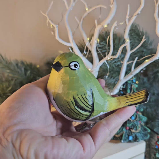 A close-up side-profile photo of a hand holding a hand-carved wooden White-Eye clip-on bird ornament, showing its lime-green crown, a distinct white eye-ring around its dark eye, carved feather lines painted in olive and dark green along its back and wing, a pale cream underside, and a bright yellow tail tip, set against a blurred background with a white branch structure and fairy lights.