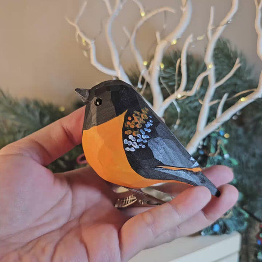 Hand-carved faceted wooden Baltimore Oriole ornament with vibrant orange body and black head, clipped to a snowy evergreen branch against a softly blurred holiday background.