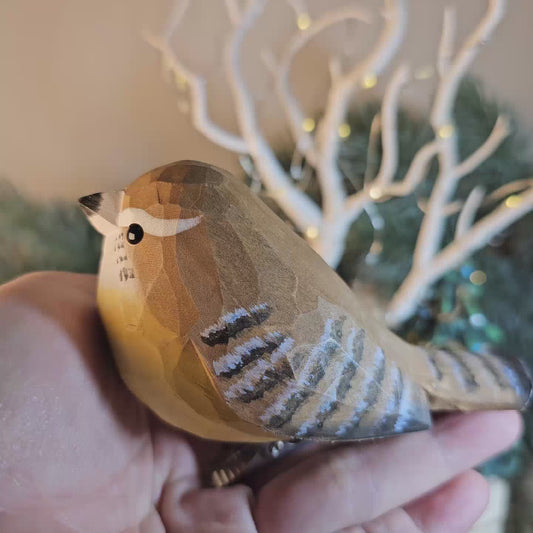 Close-up of a hand-carved, hand-painted wooden Carolina Wren clip-on bird ornament held in a hand against a blurred background of a lighted miniature white holiday tree.