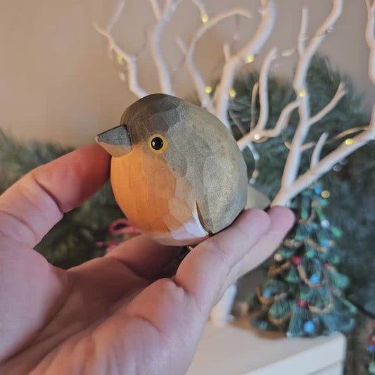 A close-up side-profile video frame of a hand-carved wooden European Robin clip-on ornament held in an open palm, featuring a warm olive-grey back and head, a bright orange-red breast, a dark eye, and a silver metal clip attached underneath, set against a blurred background with white branches and warm fairy lights.