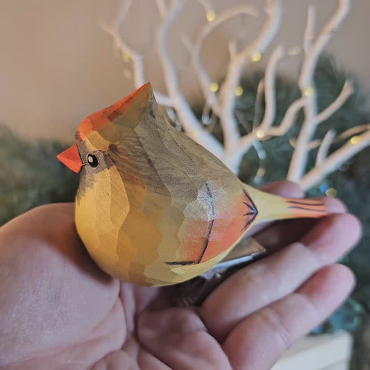 Close-up of a hand holding a hand-carved female Northern Cardinal wooden clip-on ornament, showcasing its yellow-brown body, orange beak, and metallic clip attachment against a soft-focus background.