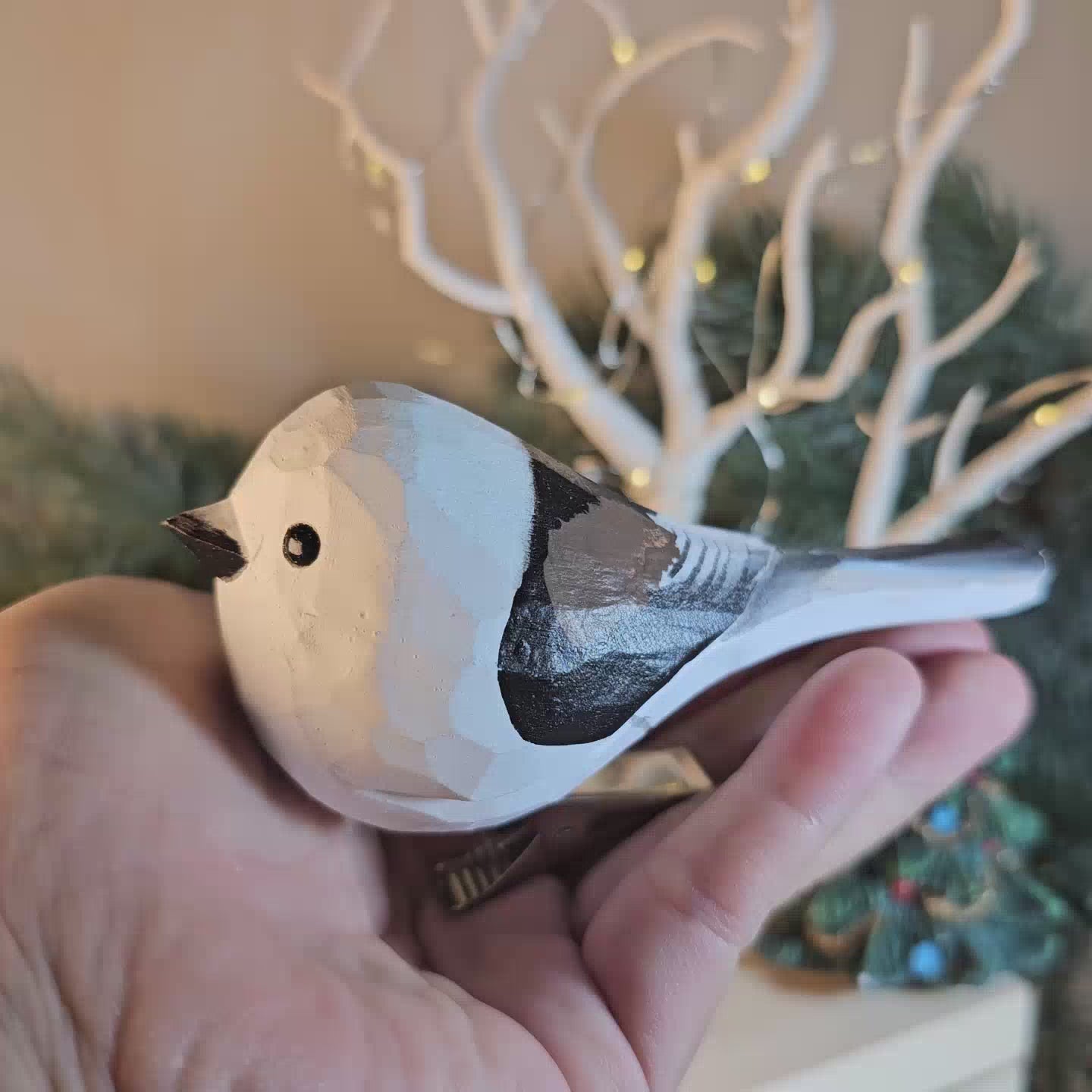 A close-up side-profile video frame of a hand holding a hand-carved wooden Long-Tailed Tit clip-on bird ornament, featuring a smooth white head and breast, a dark eye, black painted plumage along its back and wing, and a silver clip attached underneath, set against a softly blurred background with white branches and festive lights.