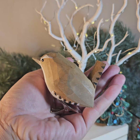 Hand-carved wooden Winter Wren ornament being held in a hand against a festive background with fairy lights and a miniature decorated tree.