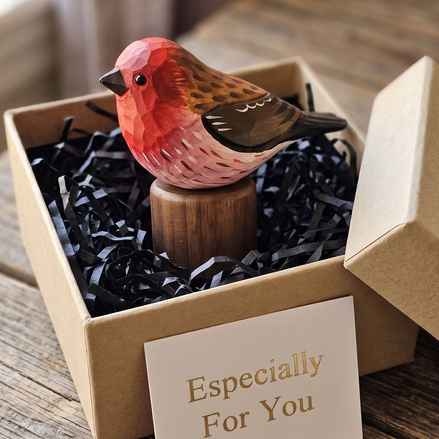A small painted Purple finch figurine perched on a wooden base, presented inside a kraft gift box with black shredded paper and a card that reads 'Especially For You,' set on a rustic wooden table.