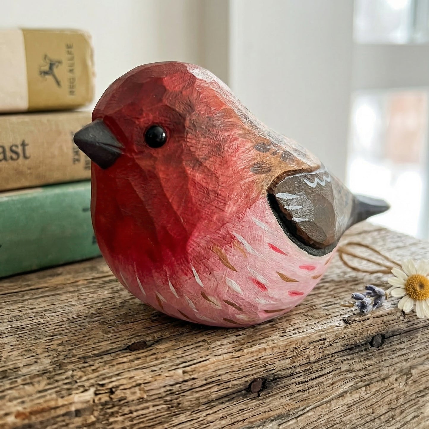 A hand-carved wooden Purple finch figurine with a red and pink body, brown wings, and a black beak, displayed on a rustic wooden surface next to old books and dried flowers.
