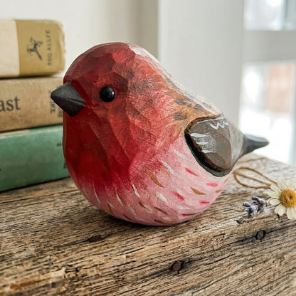 A hand-carved wooden Purple finch figurine with a red and pink body, brown wings, and a black beak, displayed on a rustic wooden surface next to old books and dried flowers.