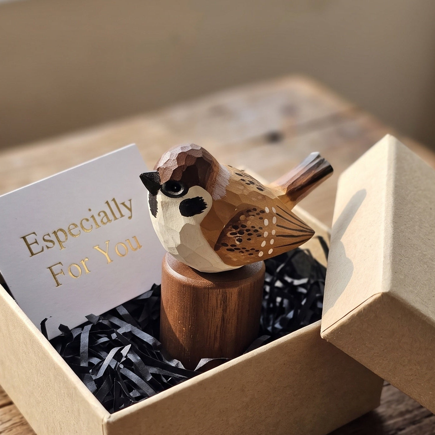 A small carved Sparrow figurine perched on a wooden stand, presented inside a kraft gift box with black shredded paper and a card that reads 'Especially For You,' set on a rustic wooden table.